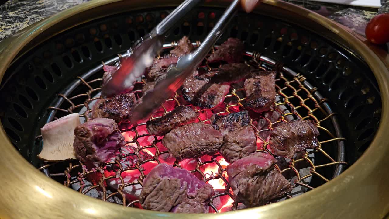 A sizzling Korean BBQ grill at Hanu Beef restaurant, chef flips juicy raw beef cube pieces of meat, pineapple, chili, and tomato, cooked over glowing charcoal for a flavorful dining experience