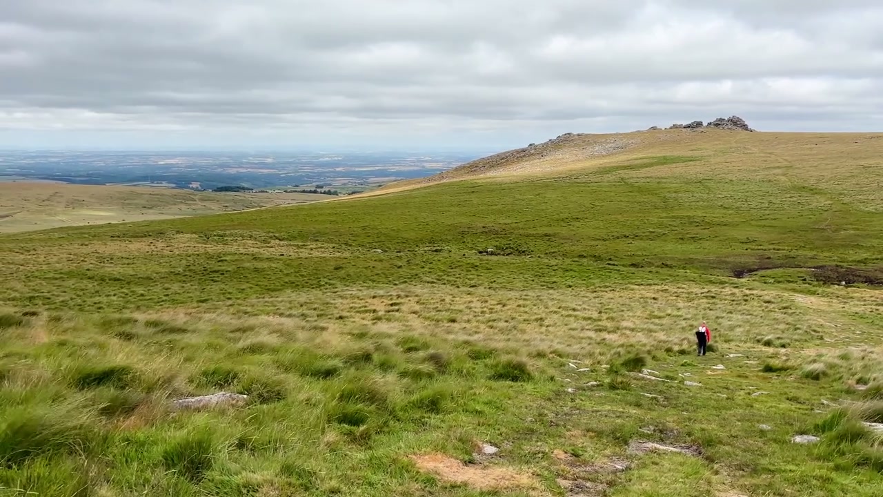 Crinale di brughiera sopra Two Bridges nel Parco Nazionale di Dartmoor, Inghilterra, con praterie ondulate, colline distanti, cielo nuvoloso e un escursionista solitario che attraversa il paesaggio.