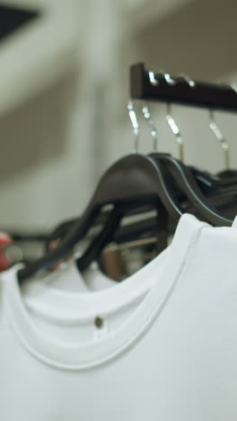hand wearing pink cloth reaches for a white top from a clothing rack, ash trouser is seen behind in a brightly lit store, with a blurred background showing other clothes neatly arranged on shelves