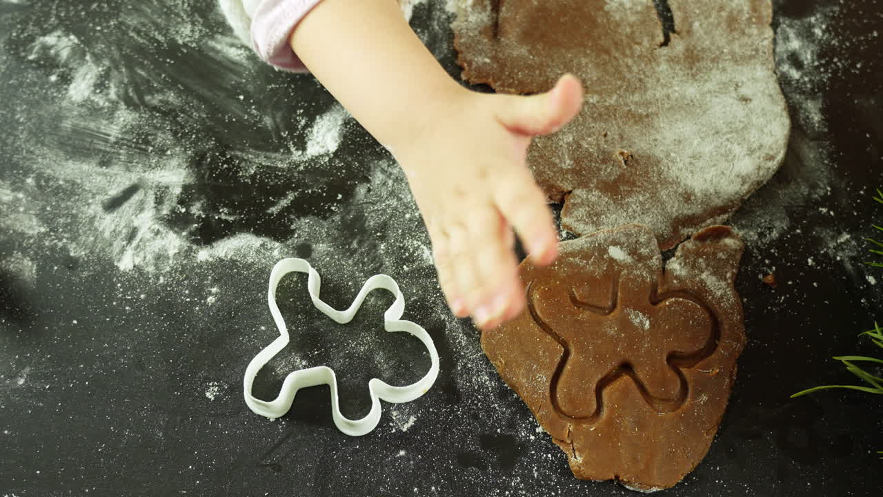 Child presses a cookie cutter into spiced gingerbread dough dusted with flour on a dark table beside a pine sprig and cutout shapes camera top-down shot, marking Christmas festive forms.