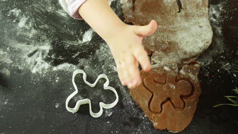 child presses a cookie cutter into spiced gingerbread dough dusted with flour on a dark table beside a pine sprig and cutout shapes camera top-down shot, marking christmas festive forms.