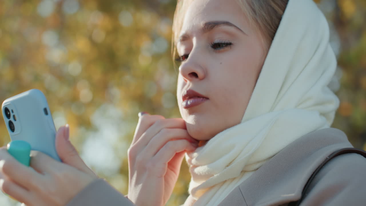 Donna caucasica all'aperto, controlla il telefono su una panchina del parco, scorre i messaggi, si sistema la sciarpa e i capelli, applica il rossetto tra un messaggio e l'altro. Atmosfera da influencer, foglie autunnali e luce solare dorata, concentrata.