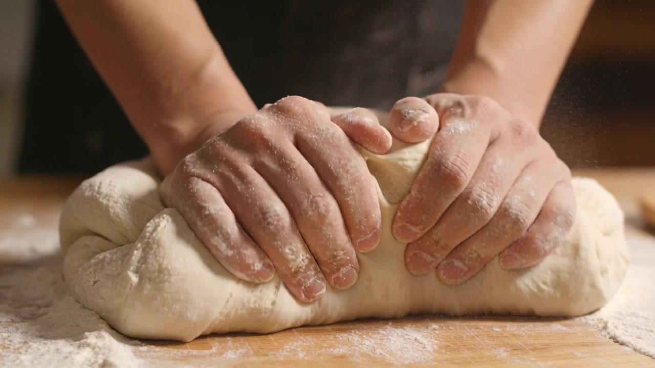 Mani infarinate che premono, piegano, pizzicano e lisciando l'impasto per formare una pagnotta su un tagliere di legno, con un grembiule scuro.