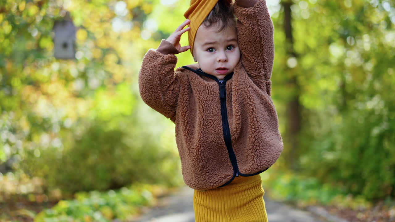 Small little kid wearing brown fluffy jacket. Baby boy takes off his yellow cap outdoors. Blurred backdrop.