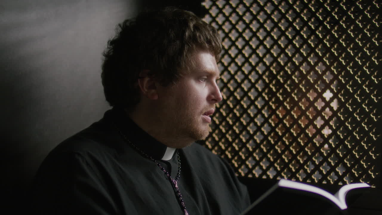 Sacerdote seduto in un confessionale che legge le Scritture dalla Bibbia