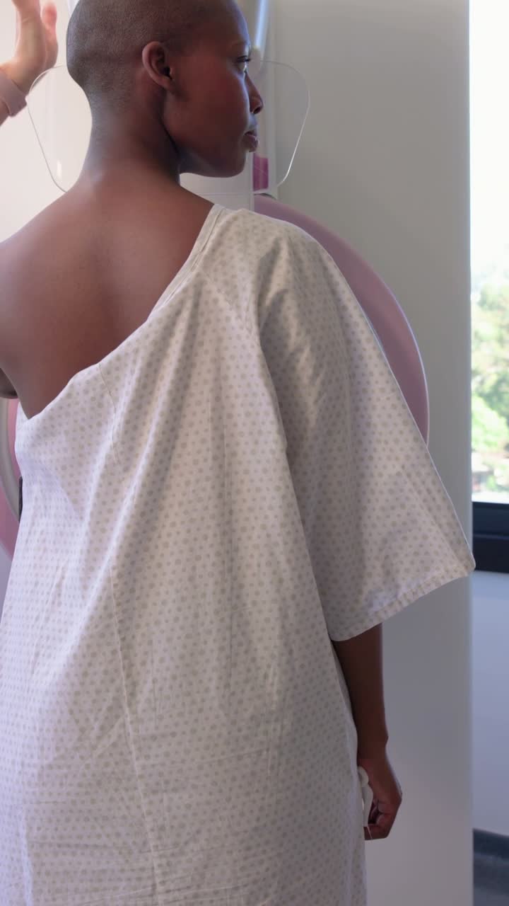 Video verticale: Donna in camice ospedaliero guidata da un tecnico in divisa sanitaria rosa che le aggiusta la spalla durante una mammografia.