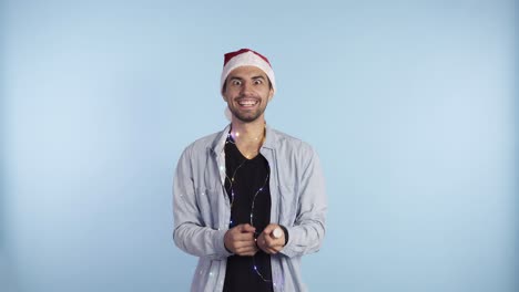 Hombre-Joven-Y-Sonriente-Con-Ropa-Informal-Y-Sombrero-De-Papá-Noel-En-La-Cabeza-Explotando-Una-Galleta-De-Confeti-Sobre-Un-Fondo-Azul-Y-Empezando-A-Saltar-De-Emoción.-Feliz-Celebración-De-Un-Nuevo-Año-O-Navidad