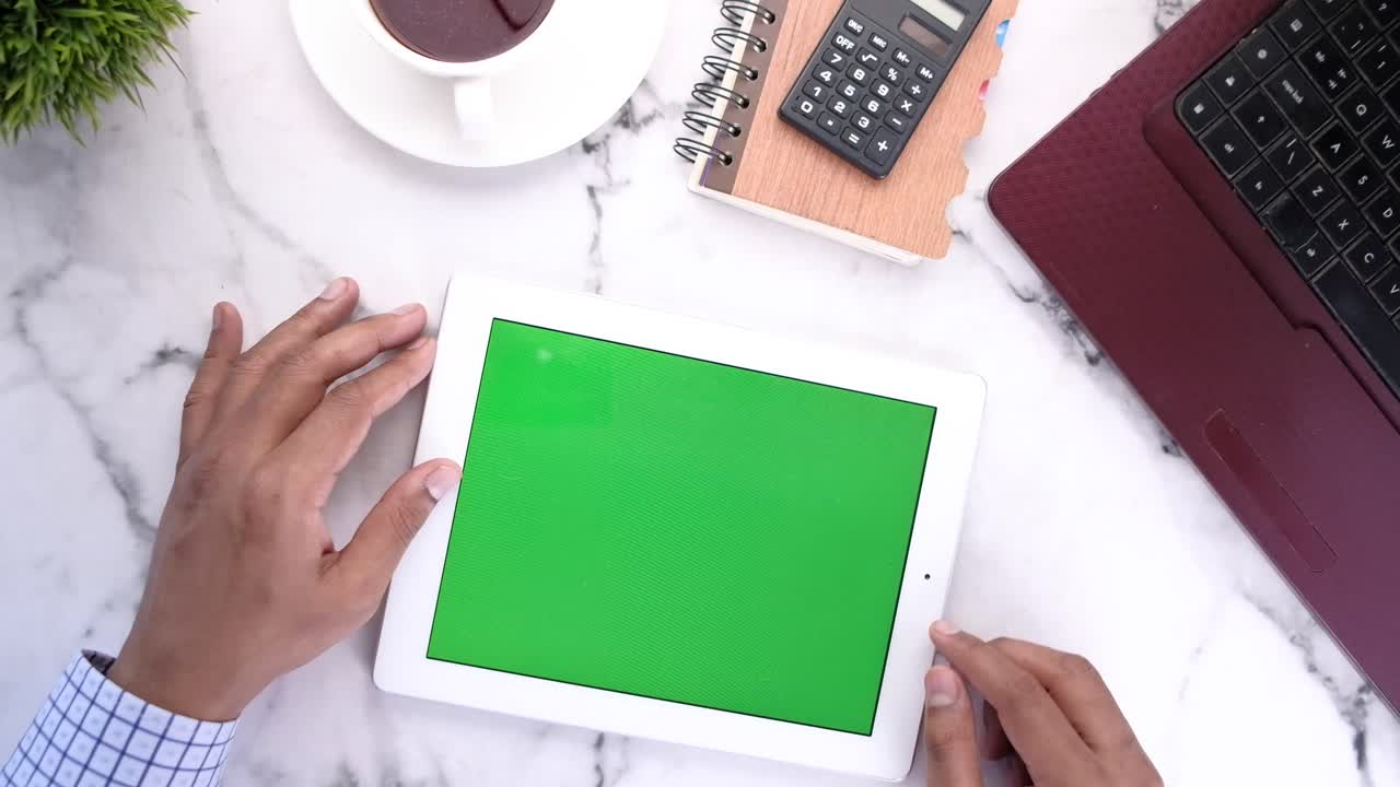 Person using tablet on a desk with laptop, coffee, calculator, and notebook