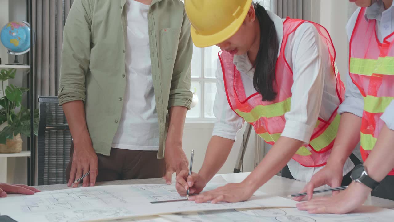 Three Asian Engineers With Helmets Helping A Man Drawing Building Construction At The Office