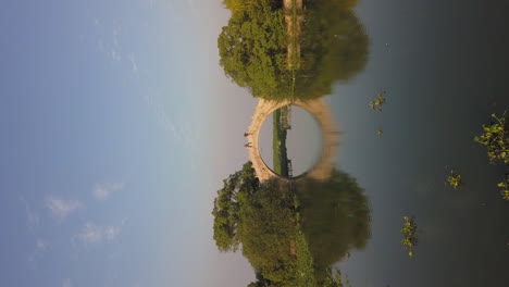 vertical format: low aerial to stone arch bridge reflected in pond