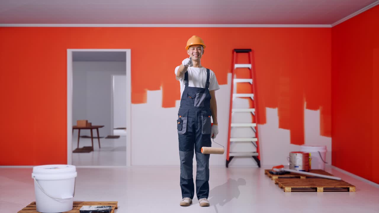 cuerpo lleno de hombre asiático pintor con casco de seguridad sonriendo y tocando su pecho luego señalando a usted mientras está de pie con la pared de pintura en la habitación