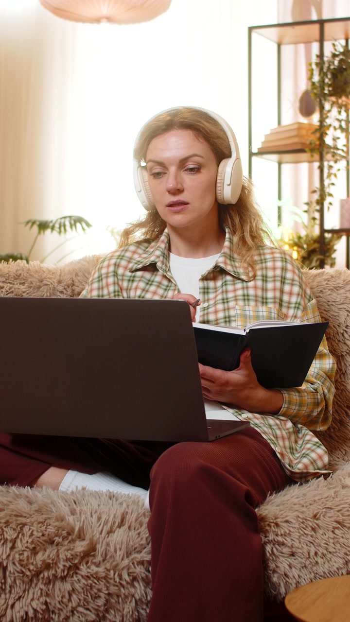 Mujer joven en sofá de casa estudiando curso en línea en portátil con auriculares y tomando notas cuidadosamente