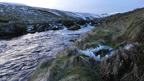 Slow-motion-footage-flowing-stream-on-an-icy-winter's-day-with-wild-grassy-hills-and-valley-in-the-background