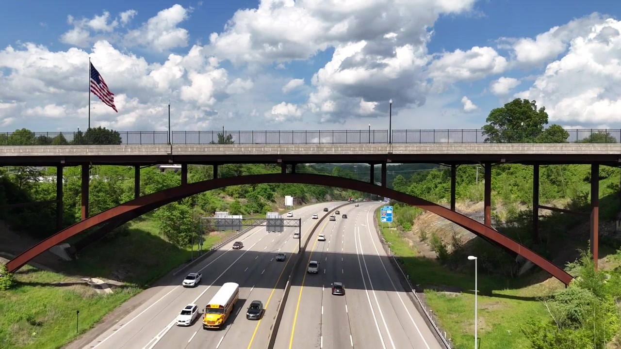 Drone Terbang di Bawah Jembatan di Atas Lalu Lintas Jalan Raya Menuju Bendera Amerika Besar