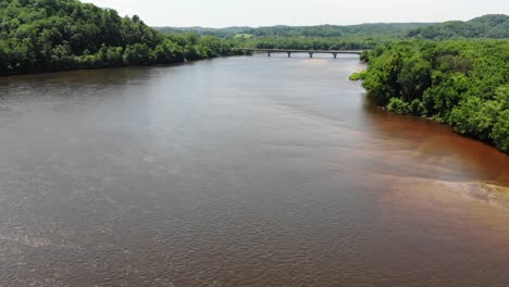 Ein-Breiter-Fluss-Im-Südlichen-Wisconsin-Fällt-Unter-Die-Autobahnbrücke,-Die-Enthüllt-Wird,-Als-Wir-Die-Kurve-Fahren,-Das-Trübe-Flusswasser,-Das-Von-Einer-Steilen-Küstenlinie-Mit-Bäumen-Umgeben-Ist