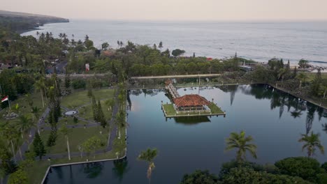 Una-Serena-Vista-Aérea-De-Una-Zona-Costera-Con-Un-Gran-Templo-Acuático-Taman-Ujung-Rodeado-De-Exuberante-Vegetación-Y-Palmeras,-Con-El-Tranquilo-Océano-Al-Fondo