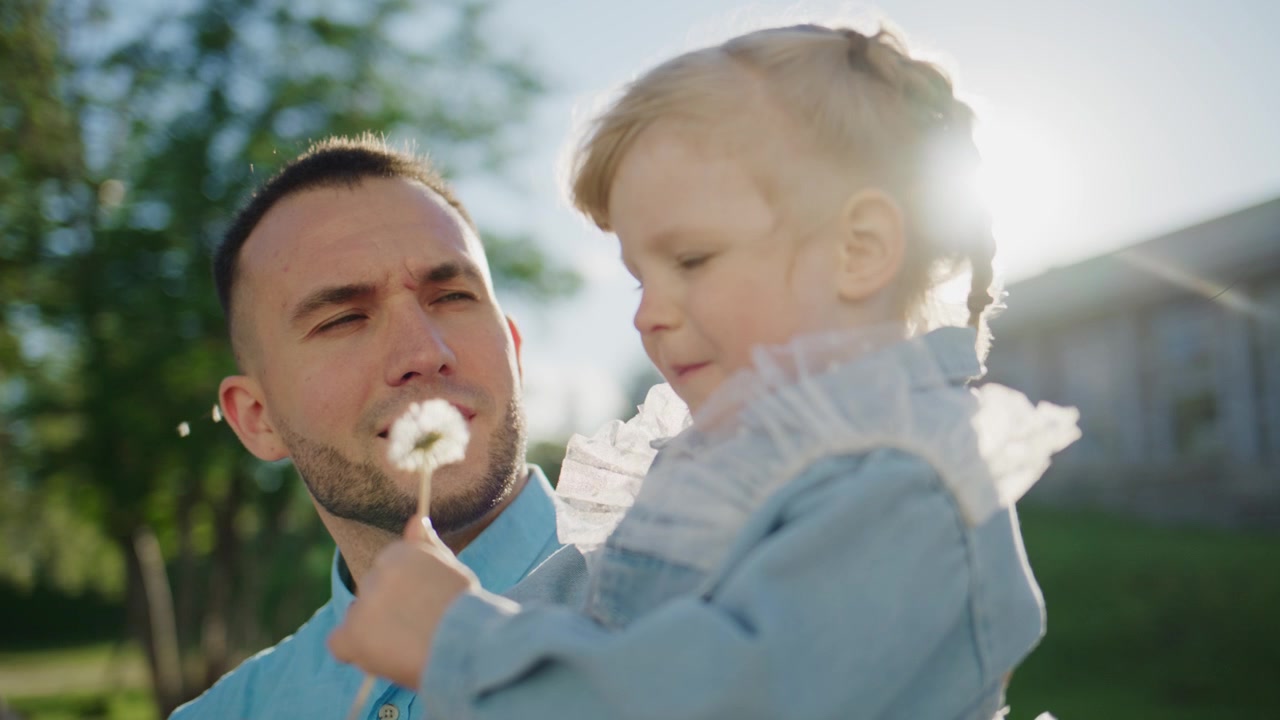 Un padre tiene in braccio la figlia piccola all'aperto. Lei soffia i semi di dente di leone verso di lui. Entrambi sorridono con dolce gioia. La luce del sole brilla dolcemente intorno a loro. Perfetto per temi familiari, primaverili o dell'infanzia