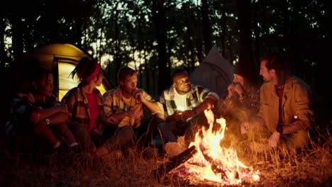 Eine-Gruppe-Von-Pfadfindern-Erzählt-Sich-Während-Einer-Rast-Gegenseitig-Gruselgeschichten-In-Der-Nähe-Eines-Feuers-Im-Nächtlichen-Wald-Mit-Zelten-Im-Hintergrund