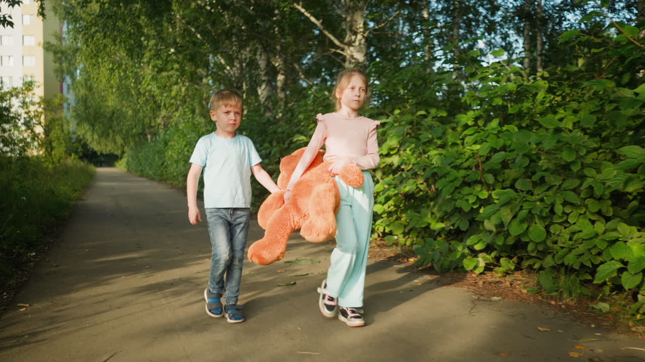 Two children walking side by side on quiet paved path lined with greenery as they carry large orange teddy bear slightly lifted off ground, under sunlight with casual expressions