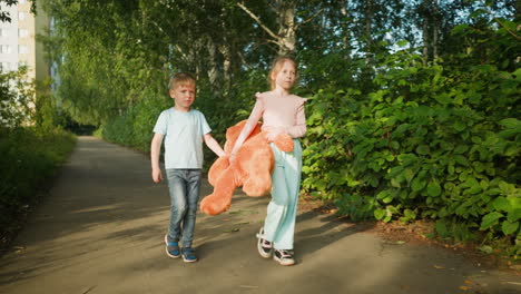 two children walking side by side on quiet paved path lined with greenery as they carry large orange teddy bear slightly lifted off ground, under sunlight with casual expressions