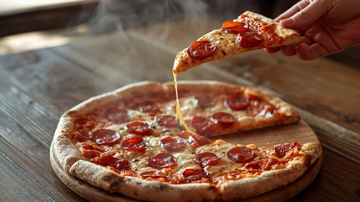 Una mano destra entra sollevando una fetta di pizza ai peperoni da un tagliere di legno rotondo, con il formaggio che si allunga.