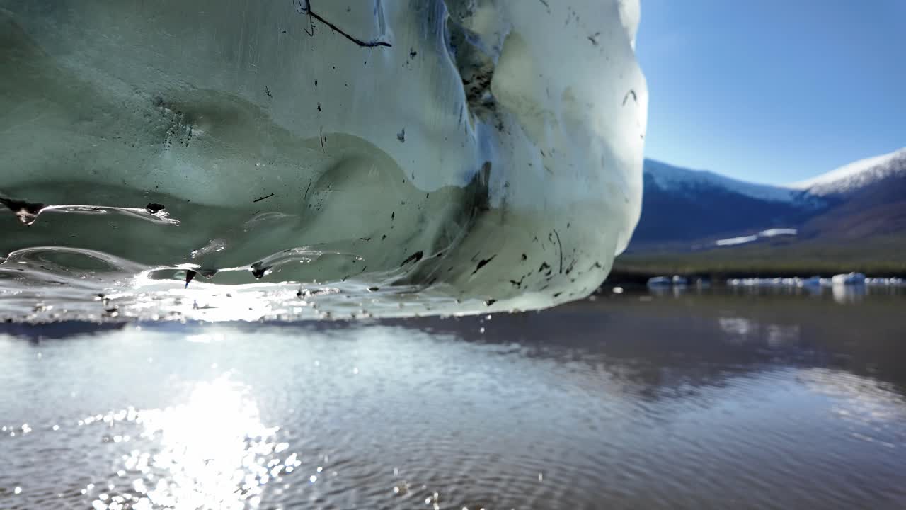 은 북극 물에서 빛 아래 녹는 빙산의 클로즈업, 멀리 눈으로 인 산과 푸른 하늘을 반영하는 평온한 물