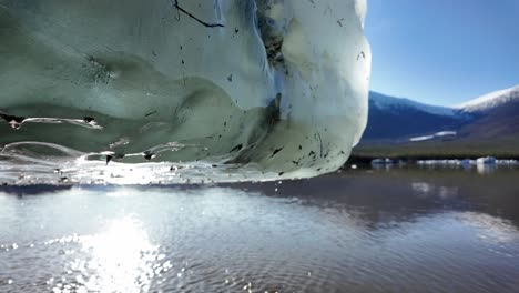 a close-up of an iceberg melting under sunlight in pristine arctic waters, with distant snow-capped mountains and calm water reflecting the blue sky