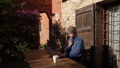 a man sitting at the laptop outside a country house, touching his goatee whilw thinking