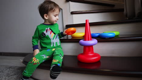 adorable kid wearing green pajama with zebras sits on the stairs at home. lovely calm baby boy building a pyramid.