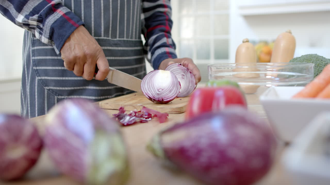 Senior african american man wearing apron cutting onion in kitchen, copy space, slow motion