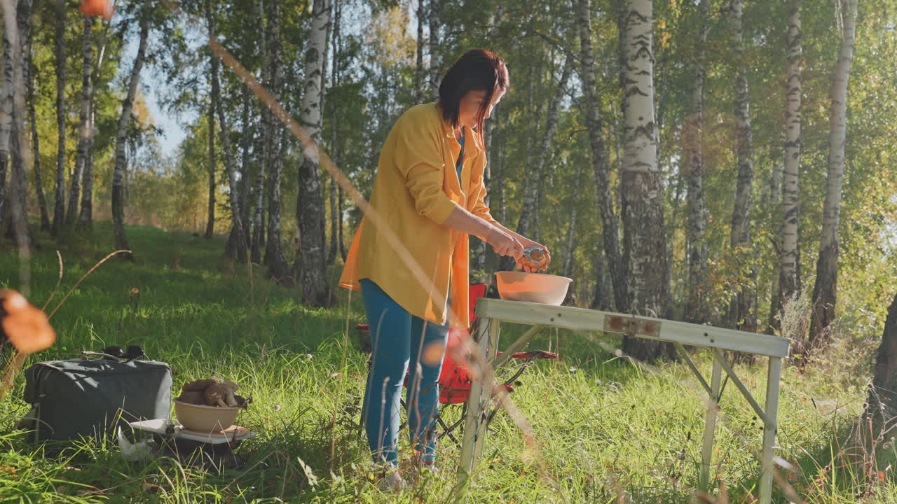 Caucasian woman preparing camping meal on portable table mixing ingredients bowl bright sunlight birch backdrop dog watching rustic cookware and fresh produce visible serene outdoor cooking scene