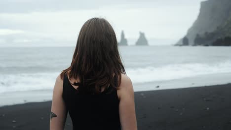 young beautiful woman in black dress standing on black sand beach iceland, looking at dramatic waves seascape, slow motion