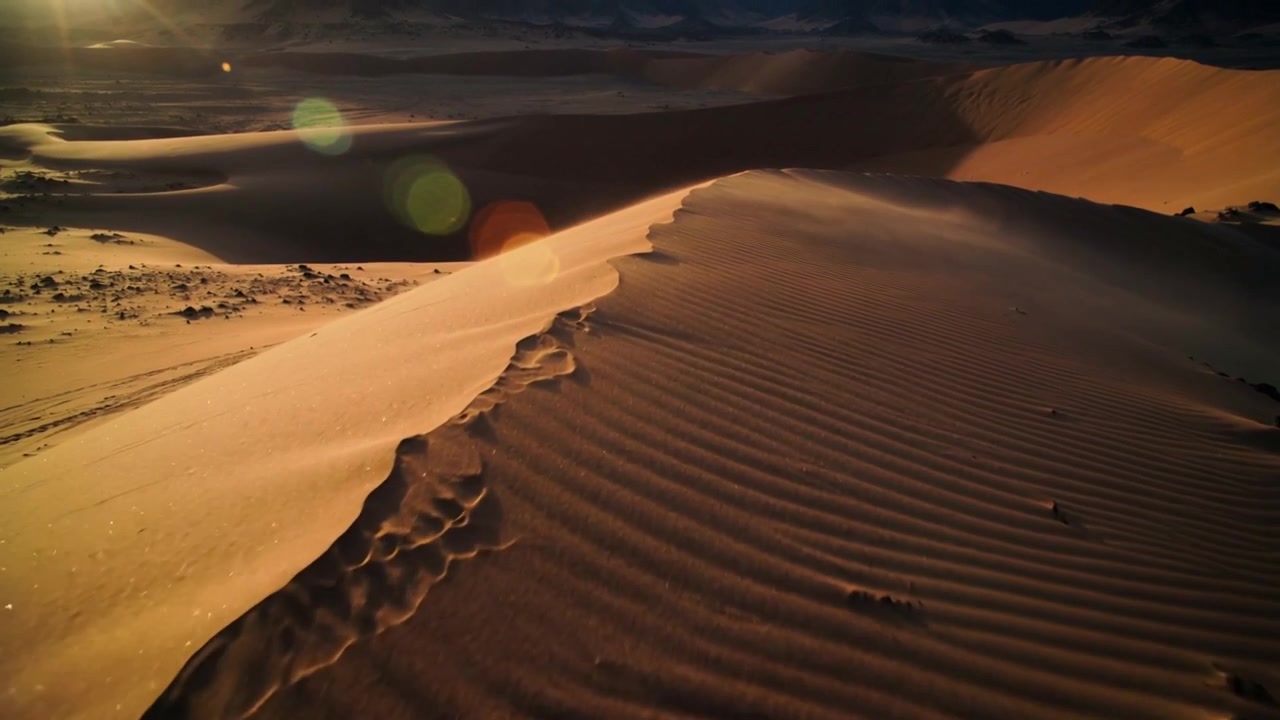 Telecamera aerea che effettua una panoramica seguendo il sole basso, rivelando una cresta di dune nel deserto con increspature, rocce e bagliore.