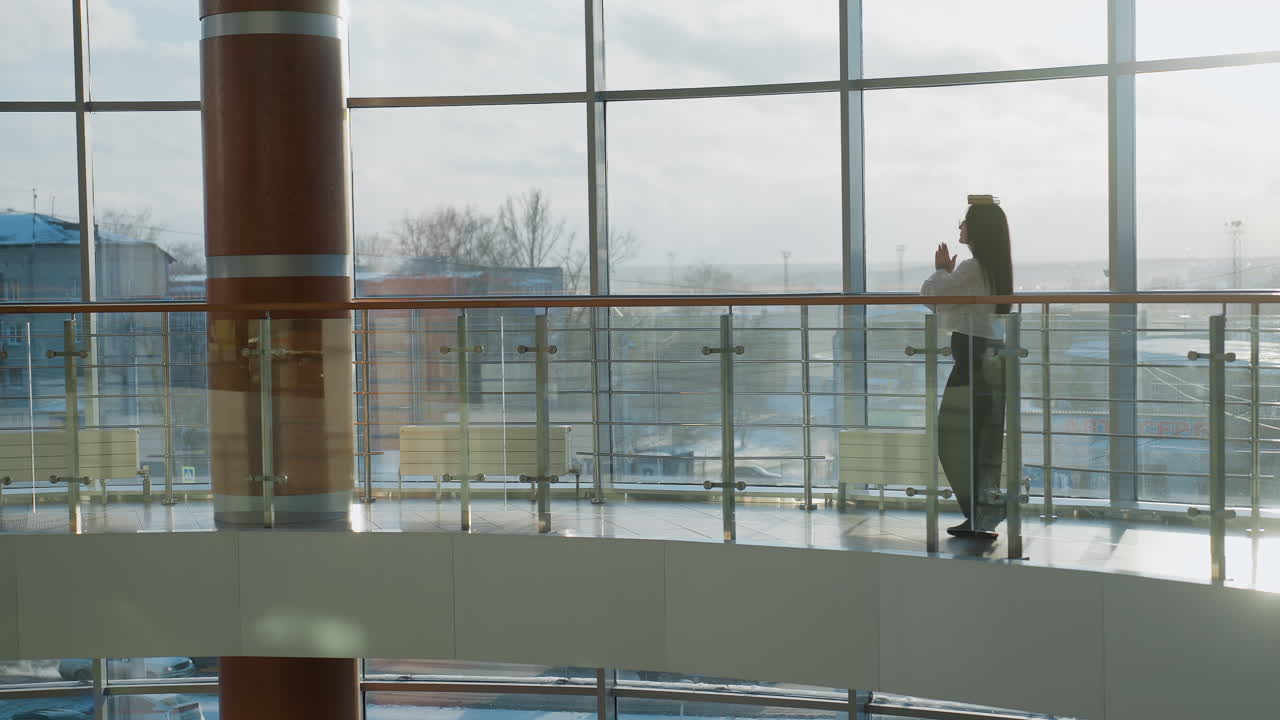 Lady walking slowly with book balanced on head along glass-paneled indoor walkway near tall wooden column, sunlit through large windows, maintaining posture and balance