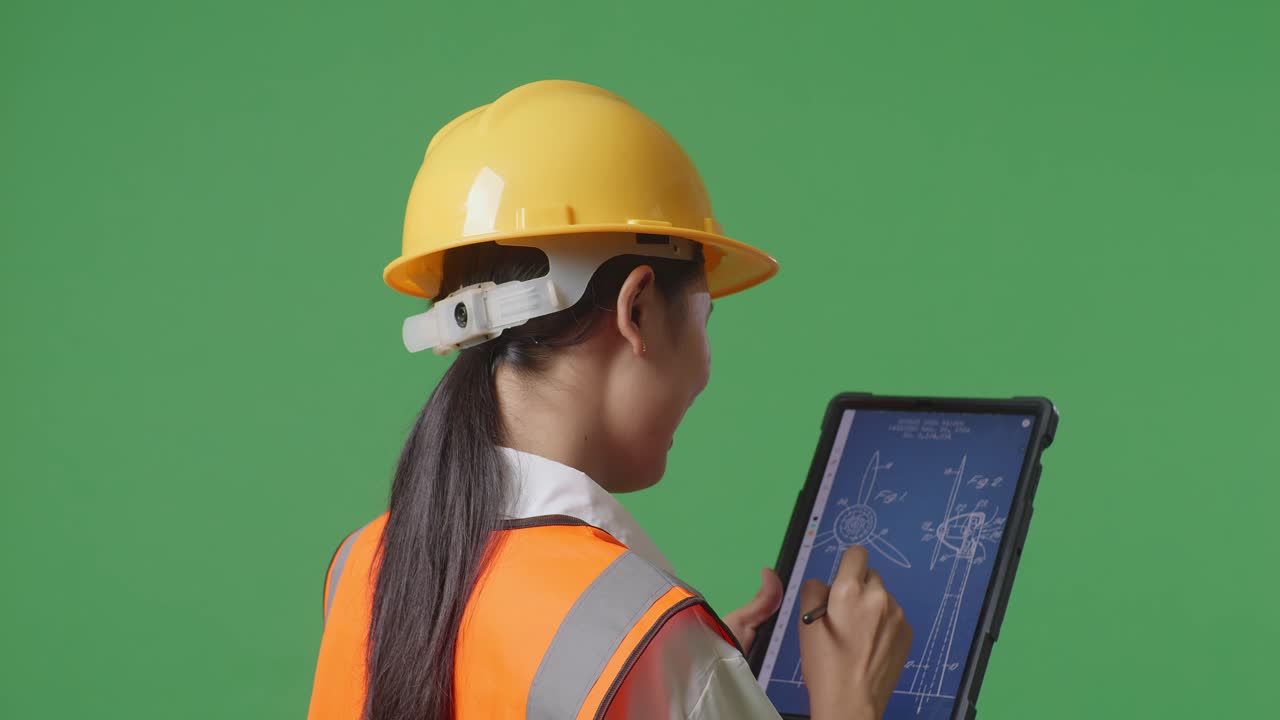 안전 헬을 착용한 아시아 여성 엔지니어가 태블릿을 사용하여 녹색 스크린 배경 스튜디오에 서서 풍력 터빈 블루프린트를 그린다.