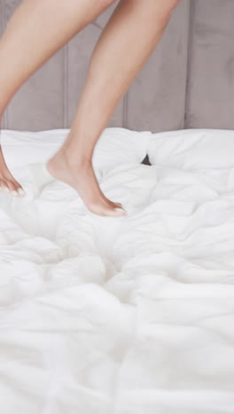 vertical video of close up of legs of biracial woman jumping on bed at home, slow motion