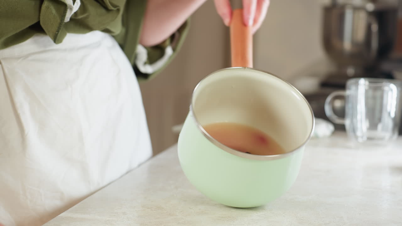 Hand view of person holding pastel green cooking pot by wooden handle and shaking contents inside over kitchen counter, apron partially visible, motion indicating meal preparation in home kitchen