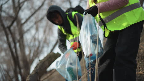 female volunteer wearing reflective safety vest and beanie holding blue plastic bag filled with collected trash during outdoor cleanup, with male volunteer in background removing litter to protect nature