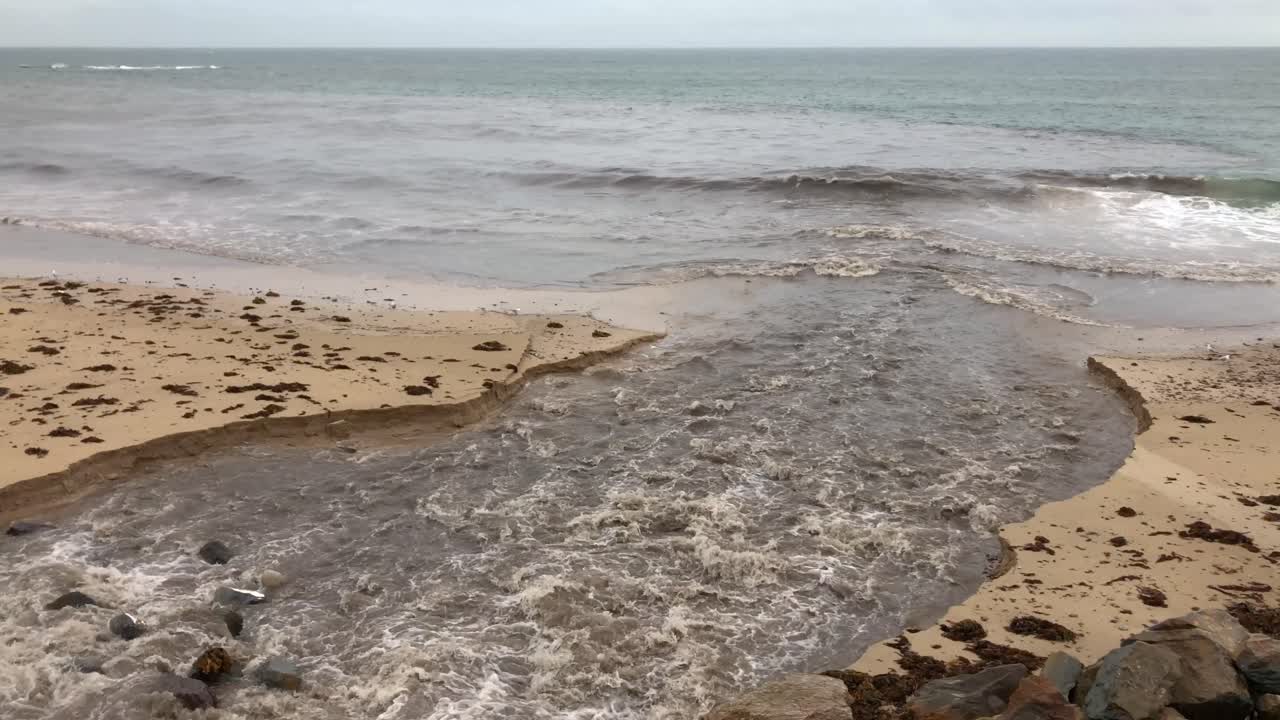 Un ruscello sfocia nell'oceano in una giornata nuvolosa, mostrando l'erosione costiera. La scena include una spiaggia sabbiosa con alghe, onde che si infrangono sulla riva e un ruscello che si fa strada verso il mare.