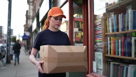 a young woman wearing an orange cap pauses outside a bookstore while holding a large cardboard box, suggesting a delivery or an interest in reading