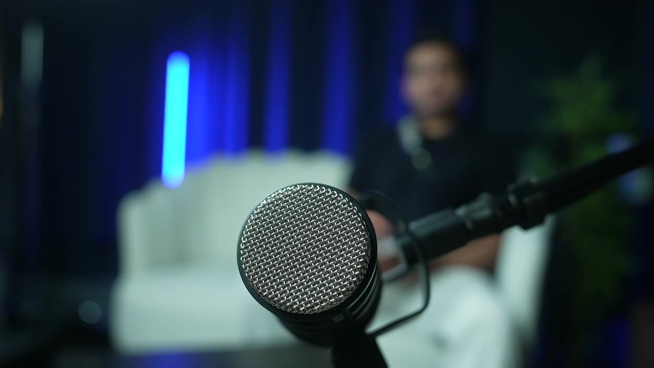 Primo piano di un microfono su asta con persona sfocata e divano in un ambiente da studio.