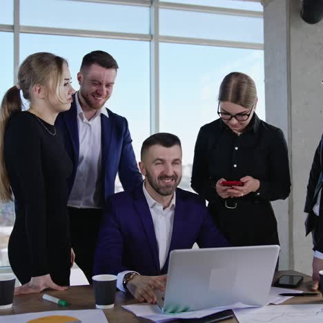 male leader sitting in front of laptop surrounded by his teammates. businesspeople communicating at work. city view in the windows of office