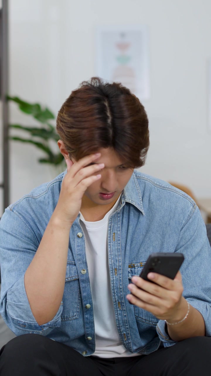 Um jovem pensativo com uma camisa jeans usa seu smartphone em uma casa moderna.