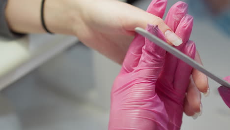 nail technician wearing pink gloves carefully files client fingernail using white opi nail file, focusing on shaping nail tip during manicure process in clean, professional salon environment