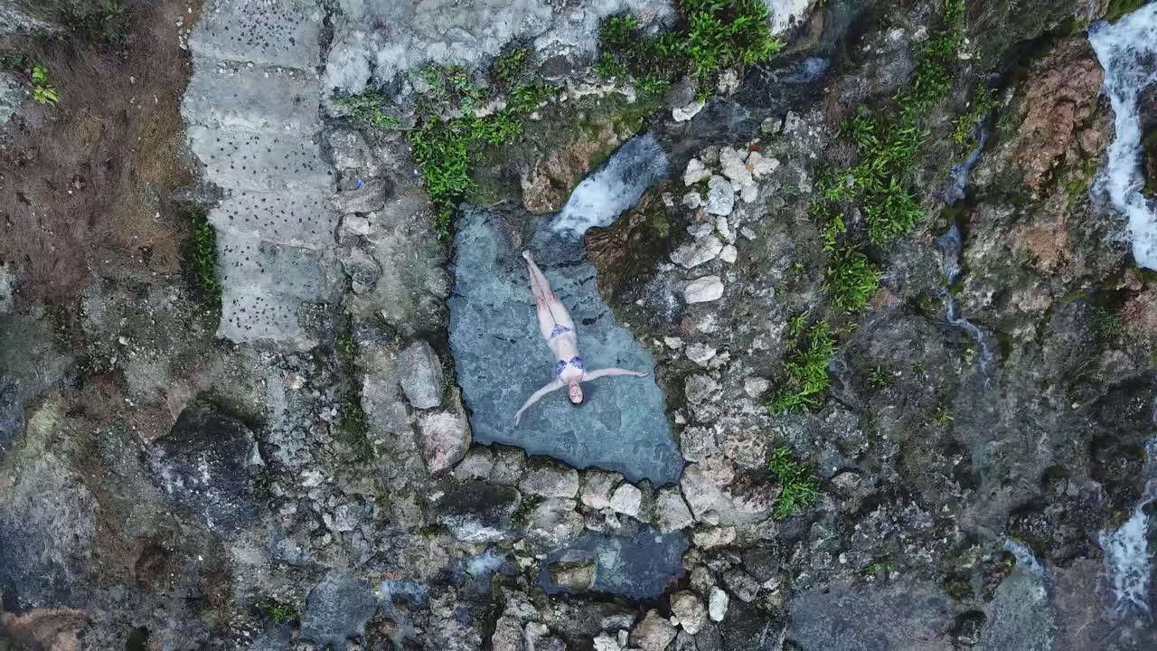 Naturally-formed pool, textured rock surface, young woman, aerial reveal
