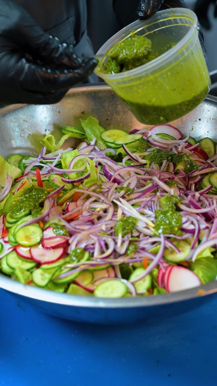 Condimento alle erbe verdi versato su un'insalata fresca di verdure. Cipolle rosse a fette e cetrioli riempiono un contenitore di metallo mentre la salsa gocciola da un cucchiaio.