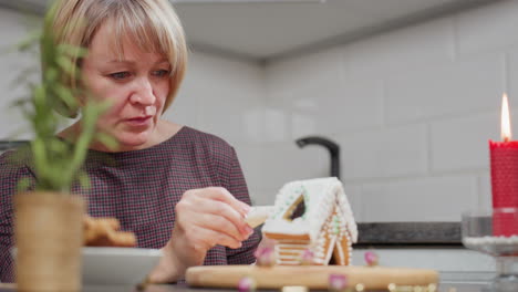 baker deeply focused on decorating gingerbread house cookie with icing, surroundings feature glowing red candle, small potted plant, and plate of cookies