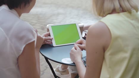 Shooting over shoulder of positive mid-adult women surfing Internet on tablet. Cheerful friends talking and looking at green screen. Modern Caucasian ladies using device with chromakey outdoors.