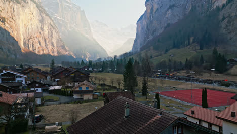 Drone-ascends-house-roofs-over-Lauterbrunnen,-Switzerland-rising-to-showcase-stunning-light-rays-passing-over-canyon-walls