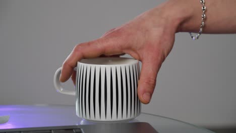 close-up of a hand picking up a modern coffee mug beside a laptop, representing daily routine, home office life, or creative work moments.